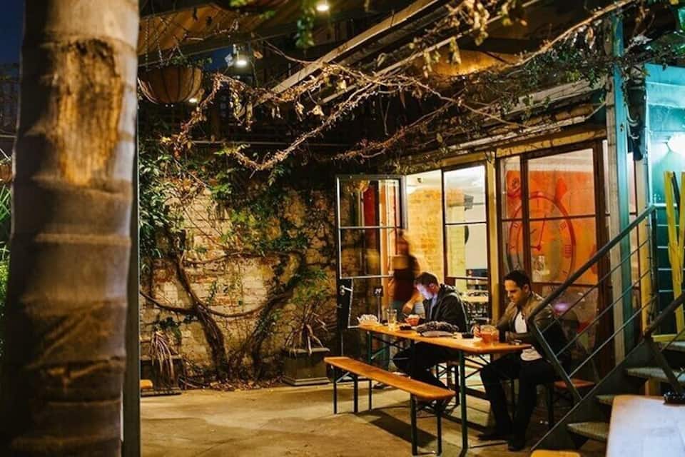 An outdoor dining area with a rustic ambiance, featuring a stone wall, wooden tables, and a staircase.