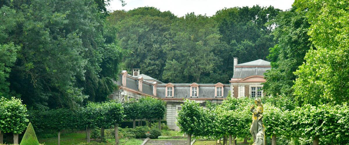 A formal garden with a statue, neatly trimmed hedges, and a large building in the background.