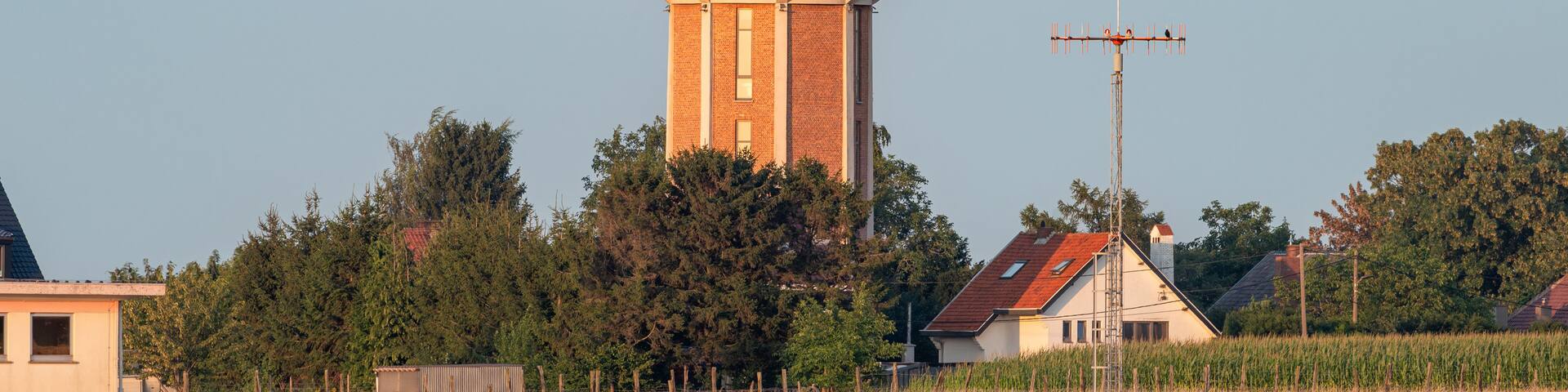 A water tower with a brick base and a metal top, surrounded by trees and houses in a rural setting.