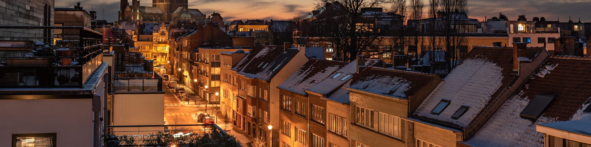 A cityscape at dusk with snow-covered rooftops, a prominent dome-shaped building, and a clear sky.