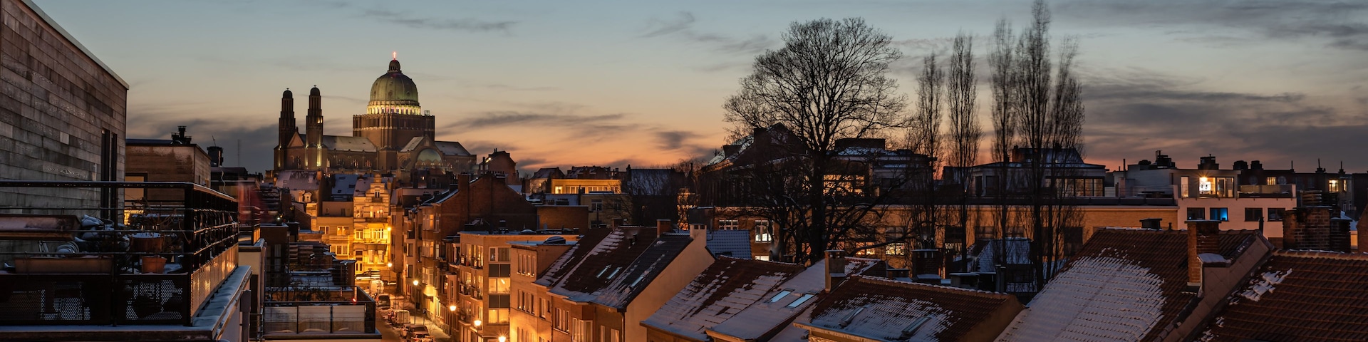 Un paisaje urbano al anochecer con tejados nevados, un edificio prominente con forma de cúpula y un cielo despejado.