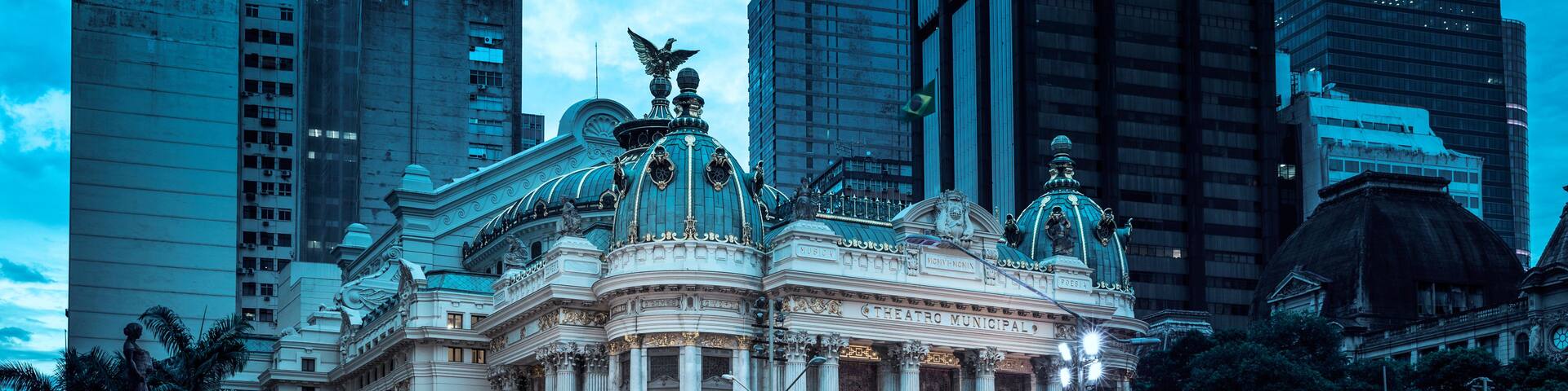 A grand, ornate building with classical architecture, including columns and decorative sculptures, set against a backdrop of modern skyscrapers.