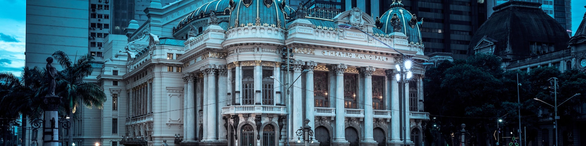 A grand, ornate building with classical architecture, including columns and decorative sculptures, set against a backdrop of modern skyscrapers.