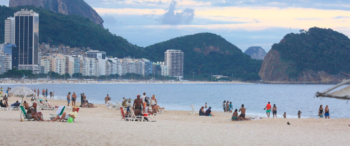 A beach with people sunbathing and swimming, a large mountain in the background, and a cityscape with tall buildings.