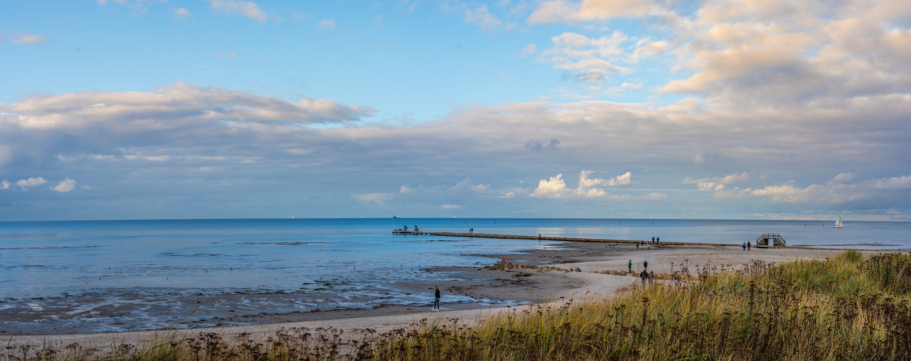A coastal landscape with a sandy beach, a pier extending into the water, and a lighthouse in the distance.