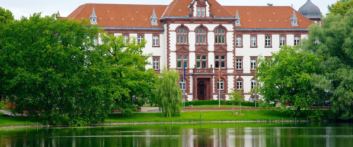 A large, historic building with a red-tiled roof and ornate architecture, surrounded by lush green trees and a calm lake.