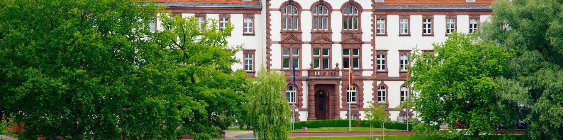 A large, historic building with a red-tiled roof and ornate architecture, surrounded by lush green trees and a calm lake.