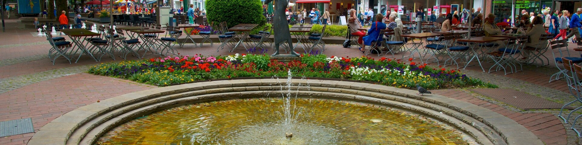 Una fuente con una estatua en el centro, rodeada de mesas y sillas bajo sombrillas azules y blancas.