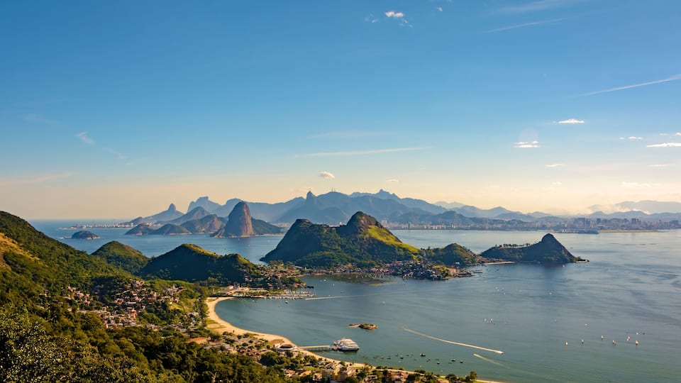 A coastal landscape with green hills, a bay with boats, and distant city skyline.
