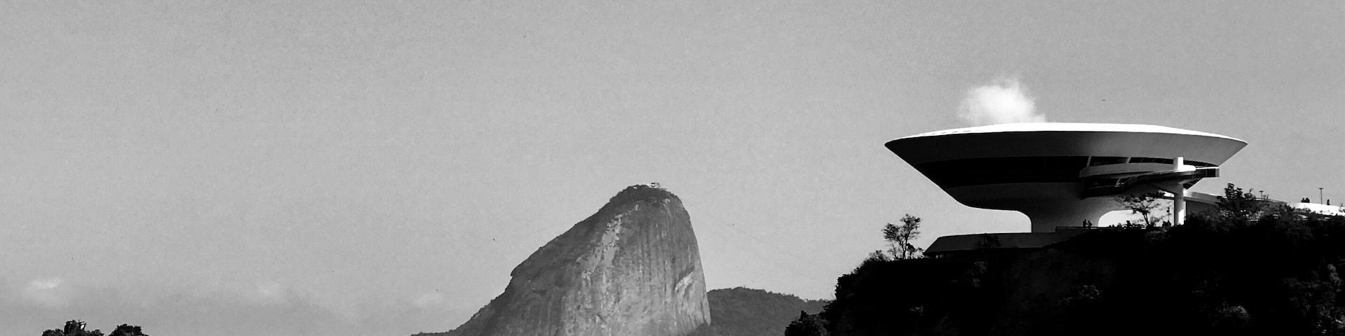 Un edificio moderno con una estructura de techo singular en una pequeña isla rodeada de agua, con una gran montaña al fondo.