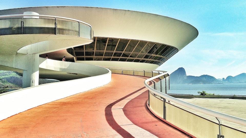 A modern architectural structure with a curved design, a pathway leading to a beach, and a mountainous landscape in the background.