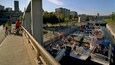 Eine Brücke mit Radfahrern, ein Kanal mit Booten und Stadtgebäude im Hintergrund.