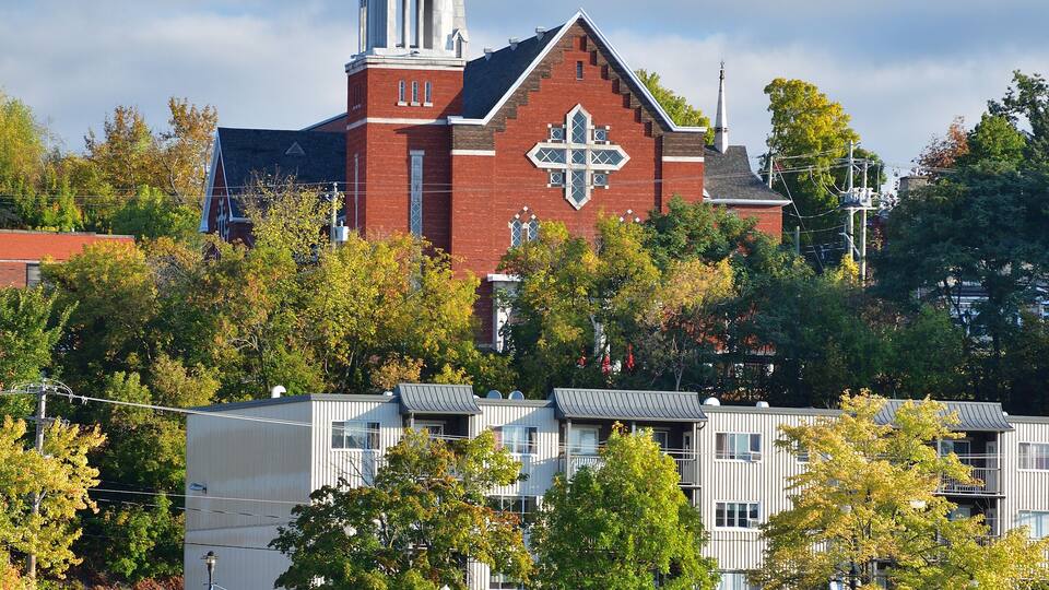 A church with a tall steeple and a cross, surrounded by trees and a body of water.