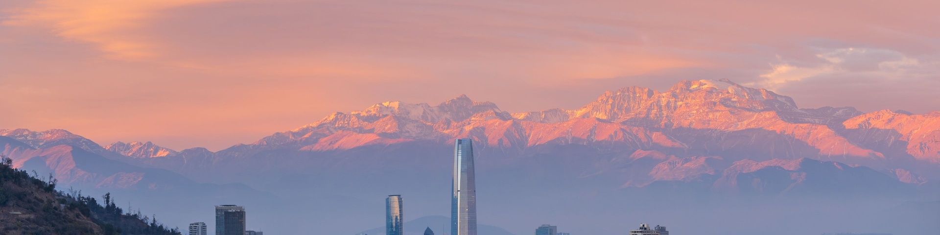 Paisagem urbana com um arranha-céu imponente, edifícios densos e uma cordilheira ao fundo.