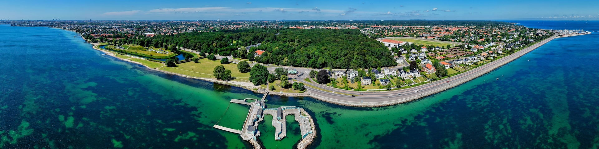An aerial view of a coastal city with a prominent peninsula, clear blue waters, and a mix of residential and commercial buildings.