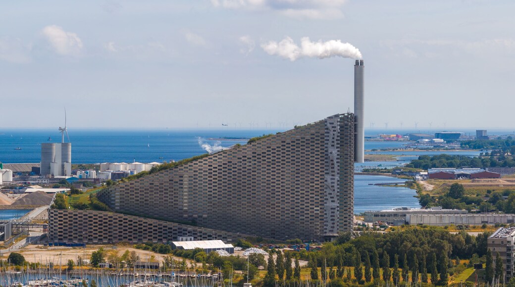Aerial view of Amager Bakke in Copenhagen, Denmark, featuring its sloped green roof, chimney, nearby wind turbines, and the Oresund Strait in the background.