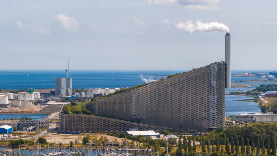 A coastal industrial area with wind turbines, a large building with a unique geometric facade, and a smokestack emitting smoke.