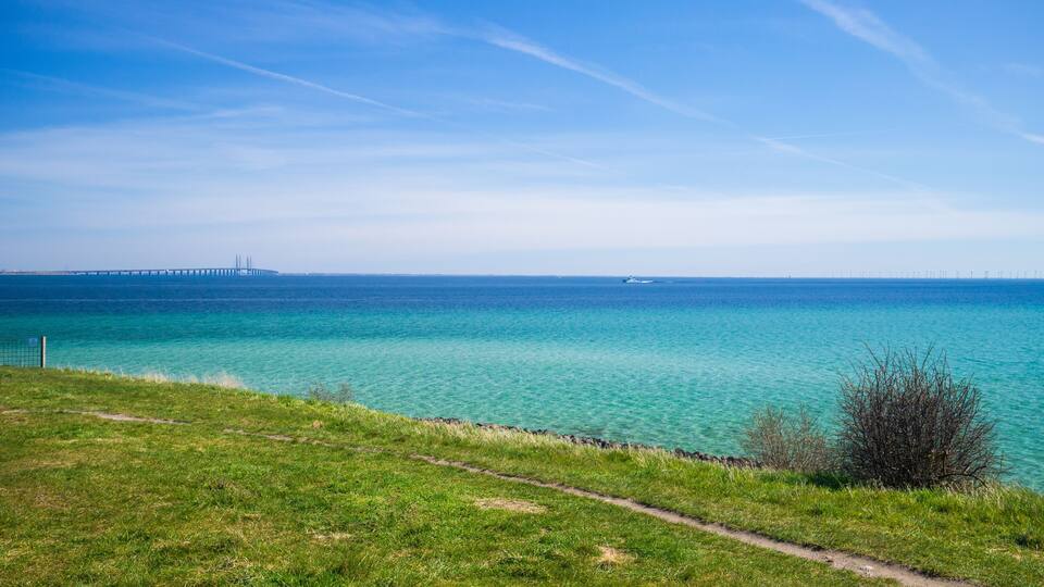 A coastal landscape with a grassy path, clear turquoise waters, and a distant bridge.