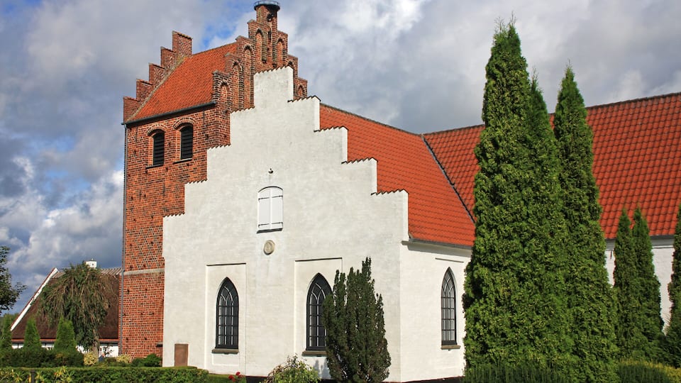 A historic building with a red-tiled roof and white walls, surrounded by well-maintained hedges and tall trees.