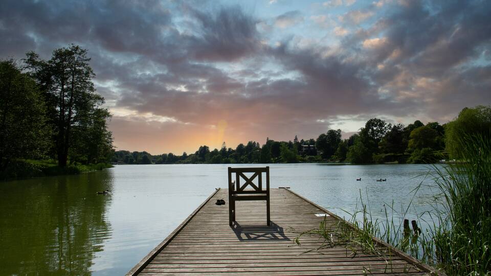 A wooden pier extending into a calm lake with a bench in the middle, surrounded by trees and a cloudy sky.
