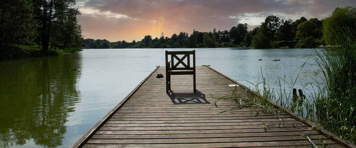 A wooden pier extending into a calm lake with a bench in the middle, surrounded by trees and a cloudy sky.