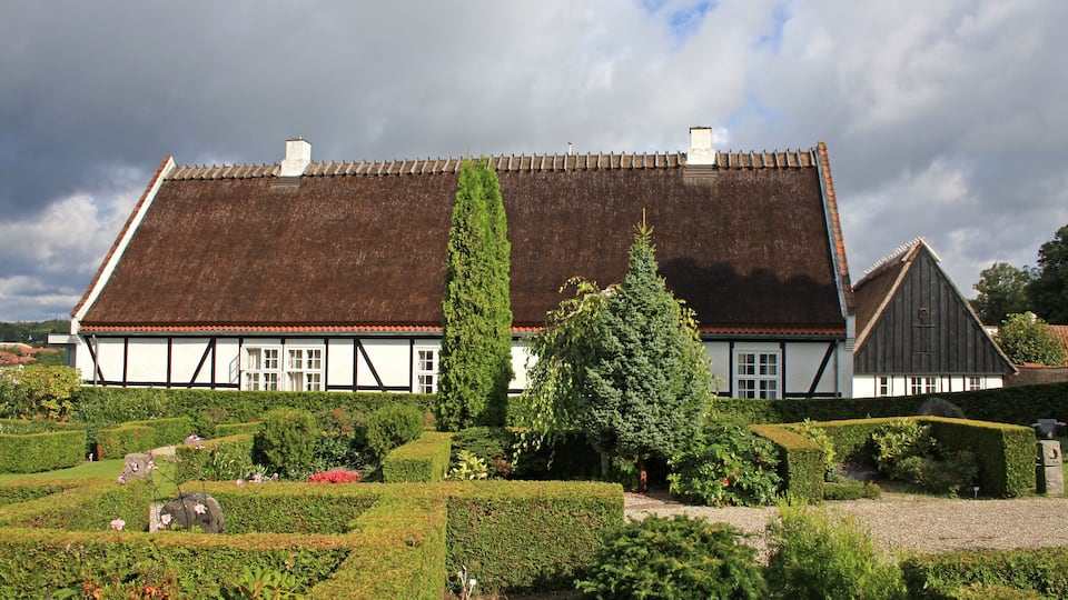 A traditional half-timbered house with a thatched roof, surrounded by well-manicured gardens and hedges.