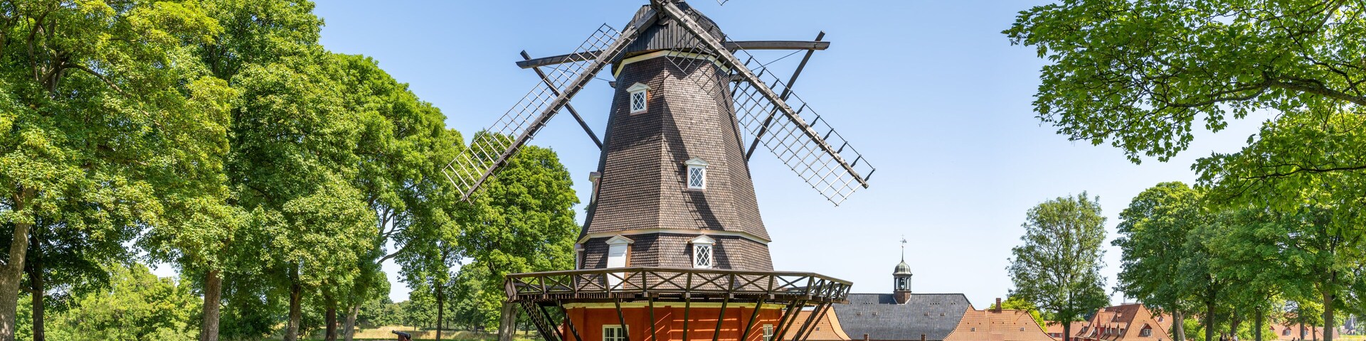 A traditional windmill with a red base, surrounded by lush green trees and a clear blue sky.