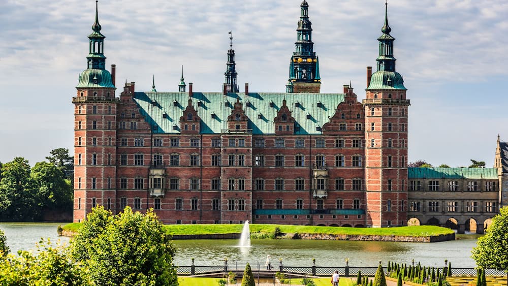 A large historic building with multiple spires, a river in front, and well-maintained gardens.