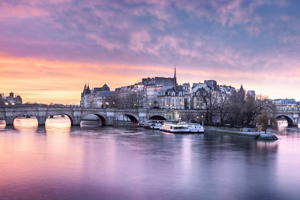 A river with a bridge and boats, surrounded by historic buildings and a sunset sky.