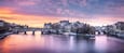 A river with a bridge and boats, surrounded by historic buildings and a sunset sky.