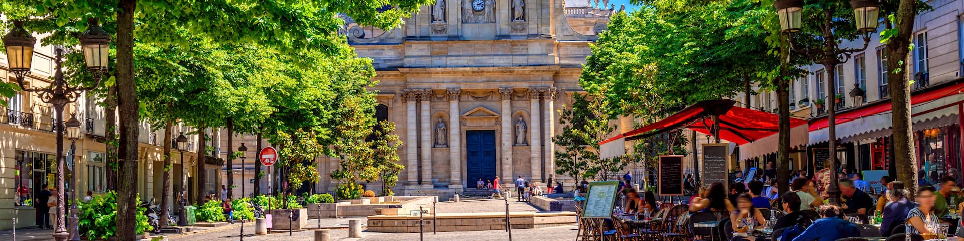 A cobblestone street lined with trees and cafes, leading to a historic building with a dome and statues.