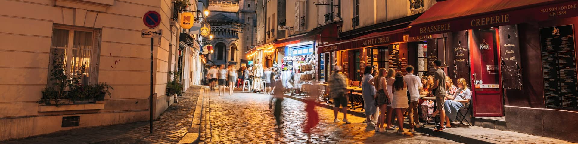 A cobblestone street lined with historic buildings, a prominent dome structure, and outdoor cafes with patrons.
