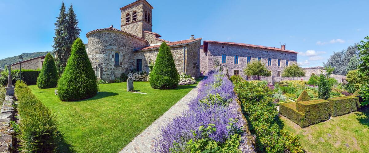 A historic stone church with a bell tower, surrounded by well-manicured gardens and cypress trees.
