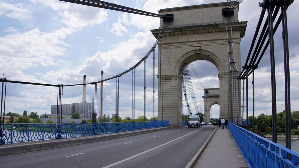 A bridge with a large arch and blue railings, a road, and a distant industrial building.
