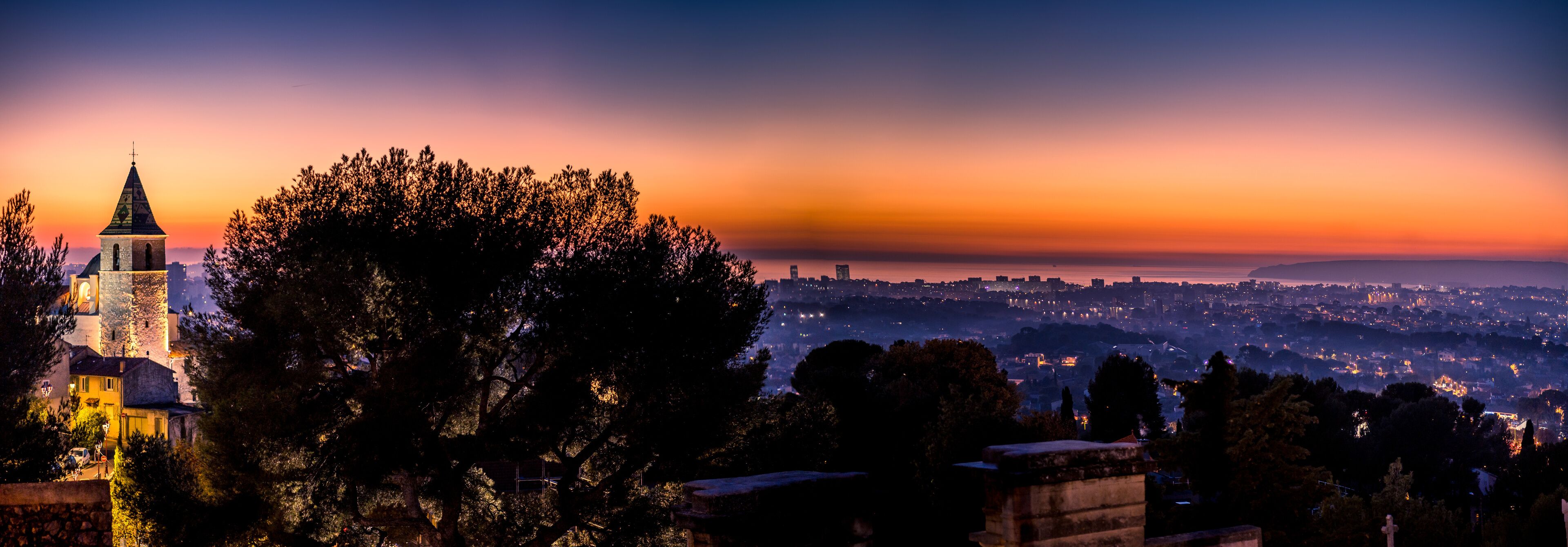 A sunset over a city with a prominent church tower and dense trees in the foreground.
