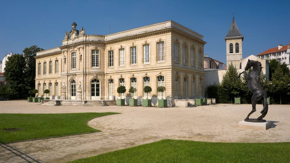 A historic building with classical architecture, a statue, and a tower in the background.