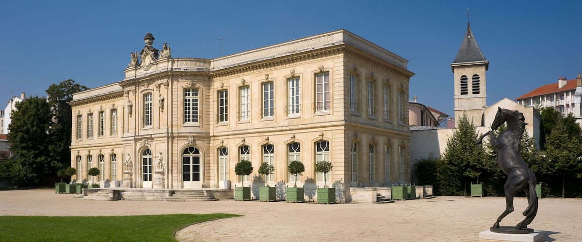 A historic building with classical architecture, a statue, and a tower in the background.