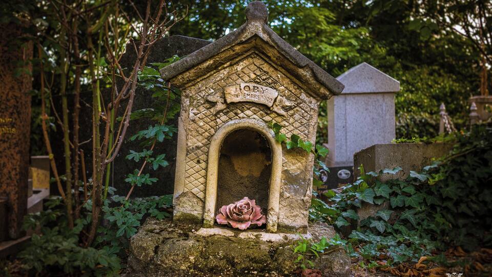 A stone birdhouse with a rose in front, surrounded by greenery.