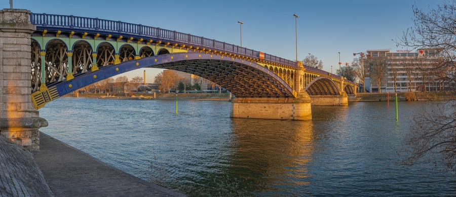 Un puente arco de acero sobre un río, con un paisaje urbano al fondo.
