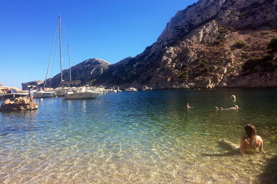 Una zona costera transparent amb vaixells amarrats prop de penya-segats i persones banyant-se a l'aigua.