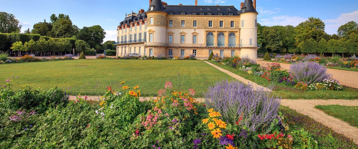 A large château with a manicured garden, a central lawn, and a variety of colorful flowers.