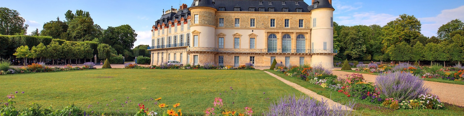 A large château with a manicured garden, a central lawn, and a variety of colorful flowers.