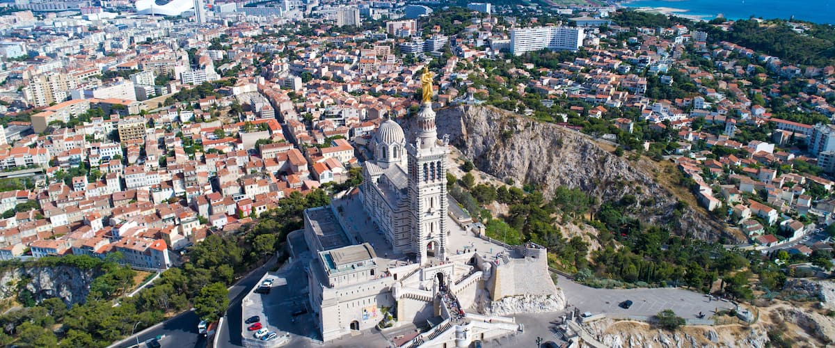 A coastal cityscape with a prominent church featuring a golden statue atop its bell tower, surrounded by residential buildings and a mountainous backdrop.