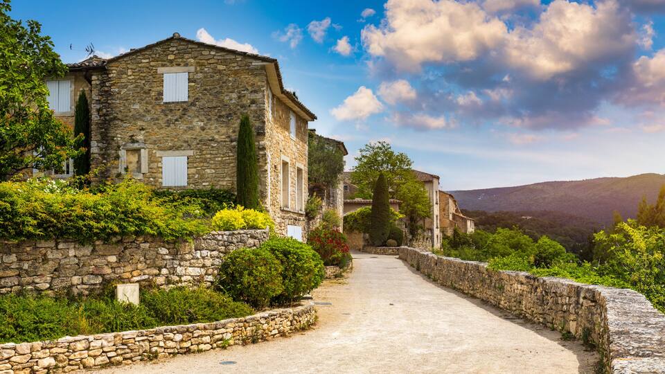 A stone building with a dirt path leading to it, surrounded by greenery and hills in the background.