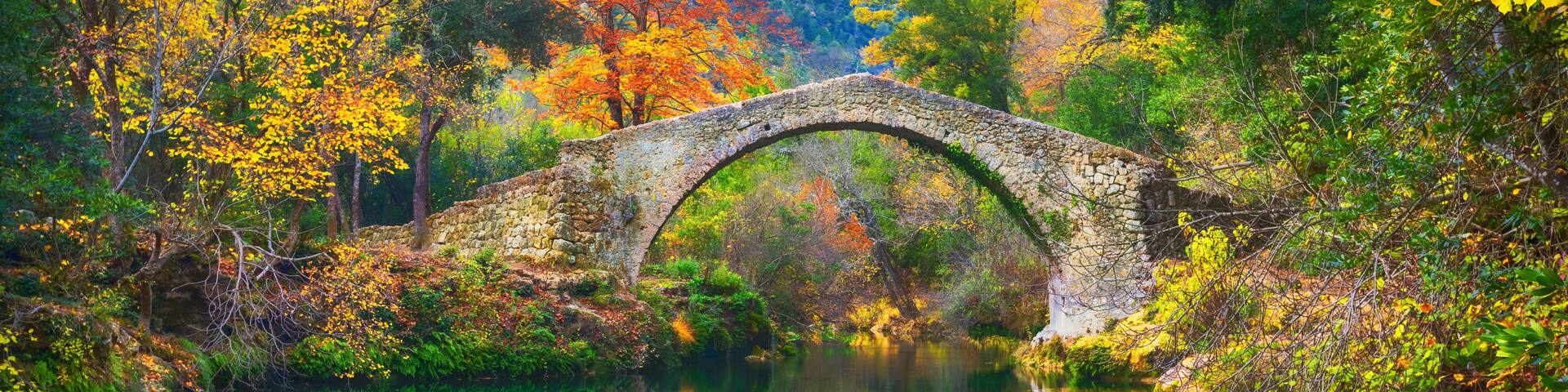 A stone bridge over a calm river, surrounded by autumn foliage.