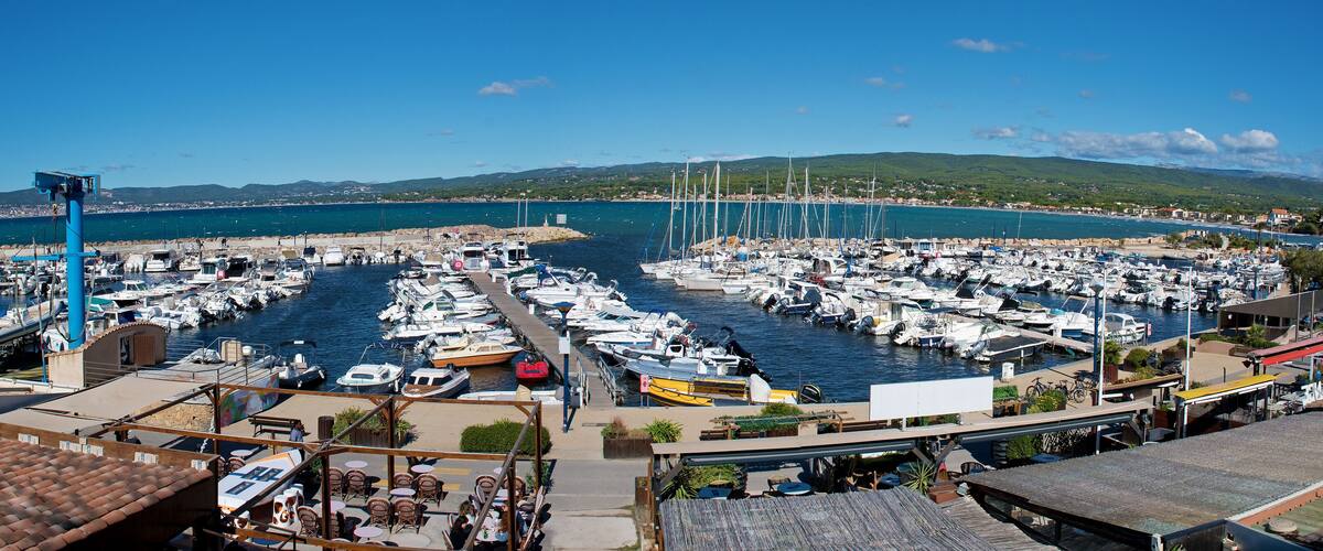 A marina with numerous boats docked, a clear blue sky, and a mountainous landscape in the background.