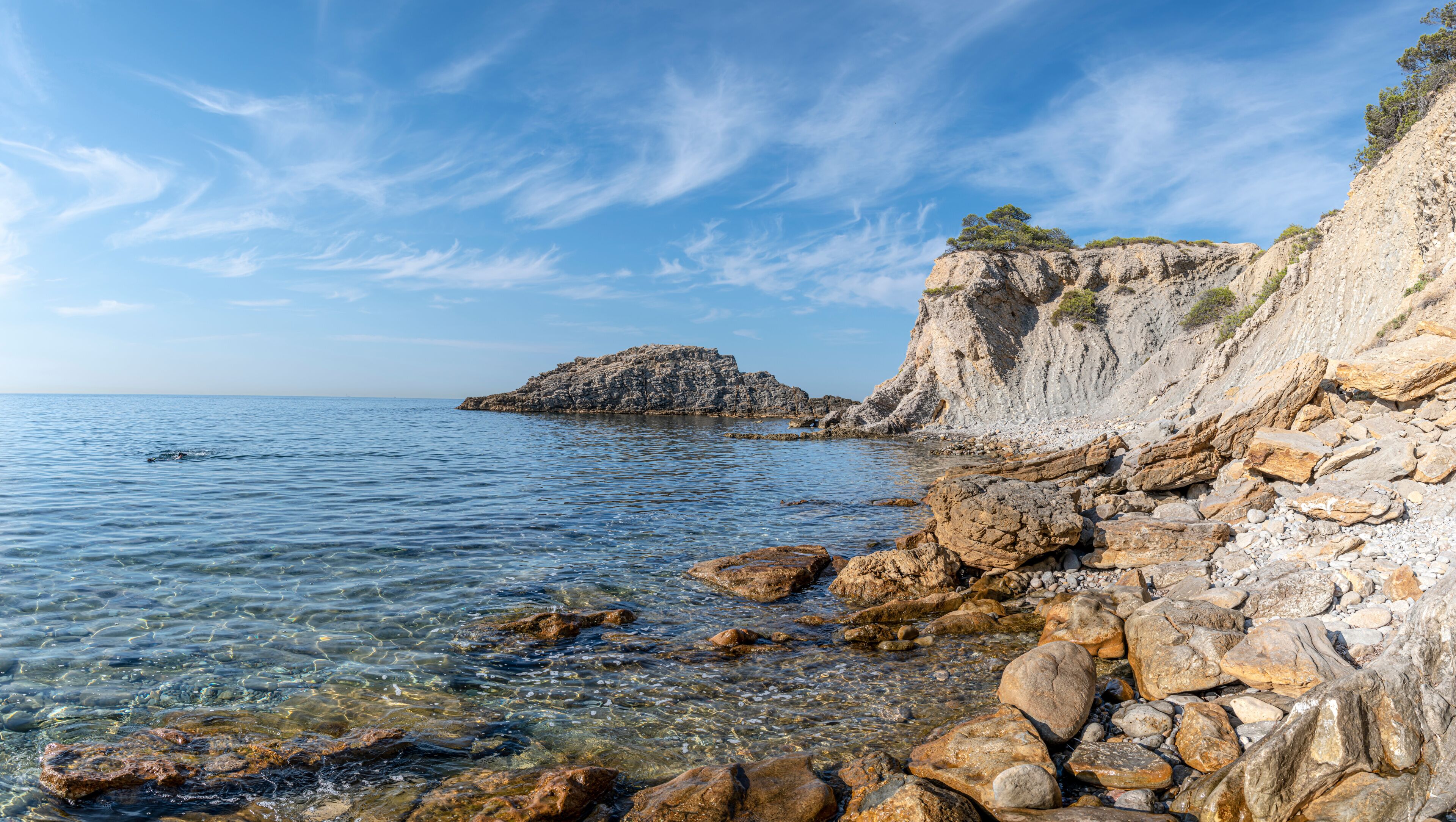 A rocky coastline with clear water, a prominent rock formation, and a cliff with sparse vegetation.