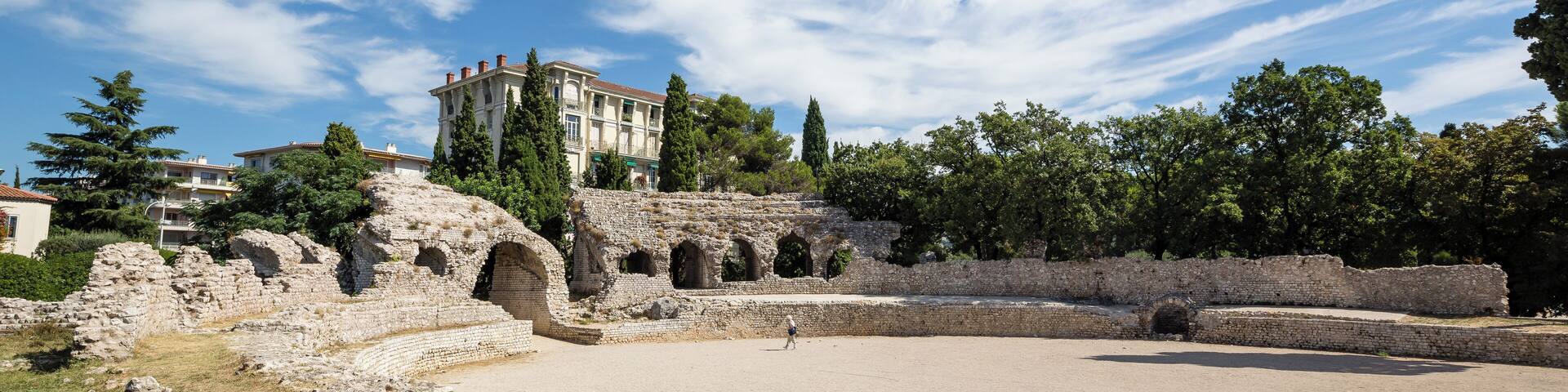 Ancient ruins with a large amphitheater, a building with columns, and trees in the background.
