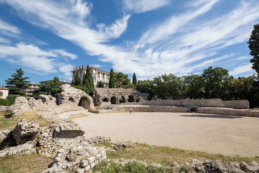 Ancient ruins with a large amphitheater, a building with columns, and trees in the background.
