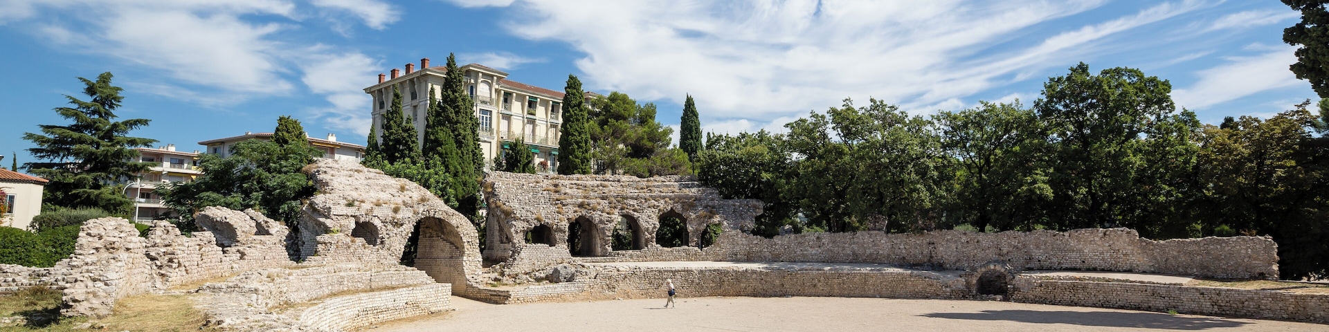 Ruinas antiguas con un gran anfiteatro, un edificio con columnas y árboles al fondo.